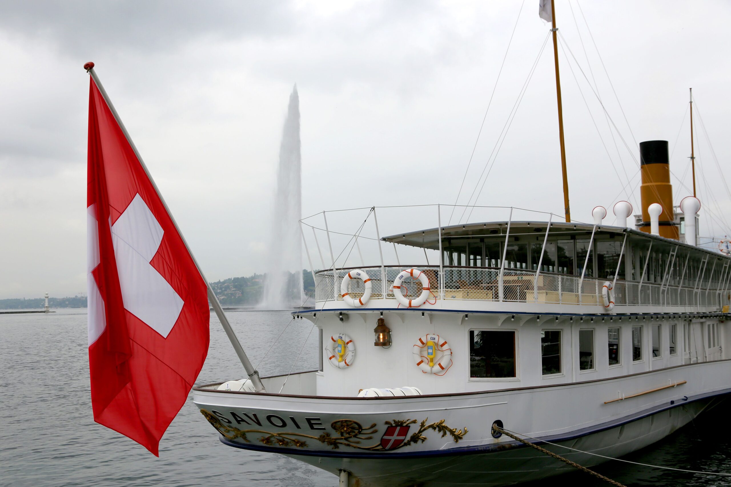 Close view of a historic paddle steamer with Swiss flag, showcasing Geneva's iconic Jet d'Eau fountain in the background.